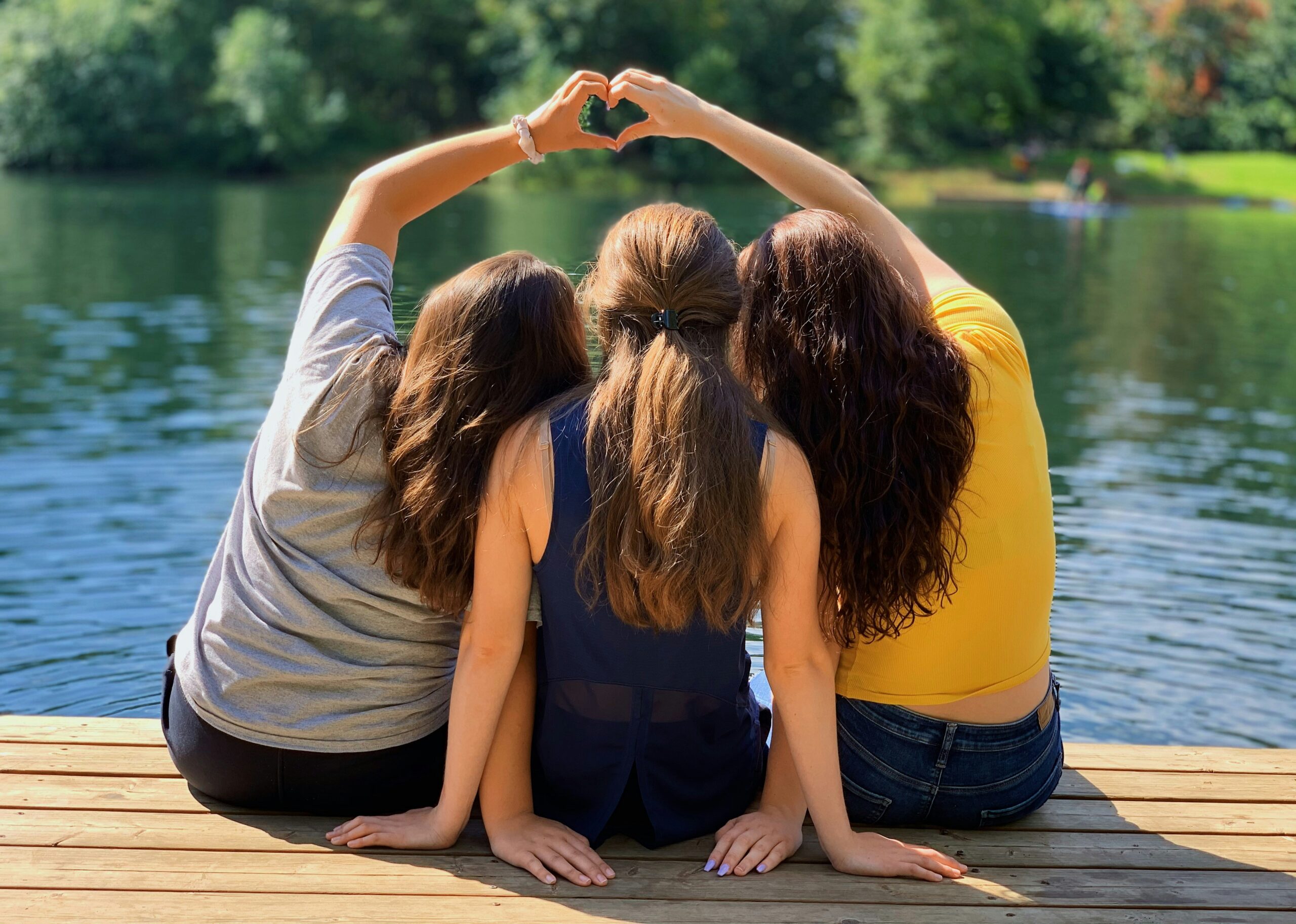 Three girls sitting by the lake and forming a heart shape with hands